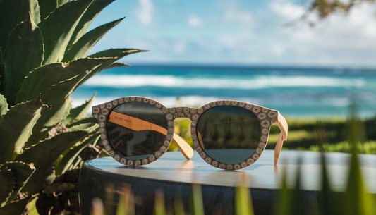 Eco-friendly sunglasses made from recycled ocean plastics and bamboo displayed in a natural outdoor setting with lush greenery, clear blue skies, and ocean waves.